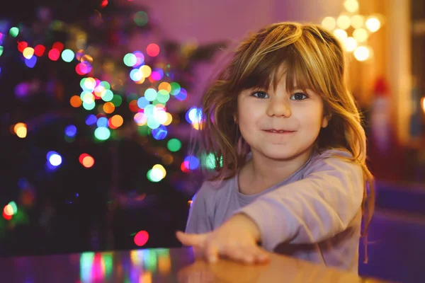 圣诞节的早上或晚上 小女孩穿着睡衣坐在那里 背景上挂着圣诞树和彩灯 健康快乐的孩子庆祝家人的传统节日 等待礼物 — 图库照片