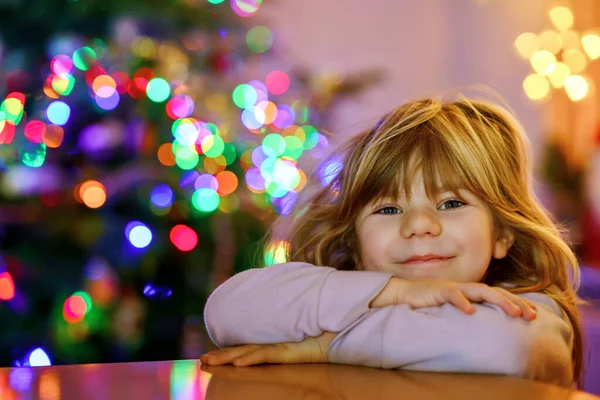 圣诞节的早上或晚上 小女孩穿着睡衣坐在那里 背景上挂着圣诞树和彩灯 健康快乐的孩子庆祝家人的传统节日 等待礼物 — 图库照片
