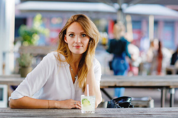 Young woman drinking mojito cocktail at cafe terrace at hot summer day. Beautiful businesswoman enjoying warm evening at afterwork restaurant. Happy smiling alone lady