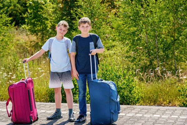 Two children, school boys with suitcases before leaving for summer vacation camp. Happy kids, siblings, twins brothers going on journey, family road trip waiting for train