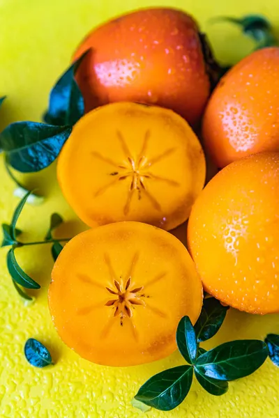 Whole and cut half persimmons with leaves and water drops on yellow background. — Stock Photo, Image