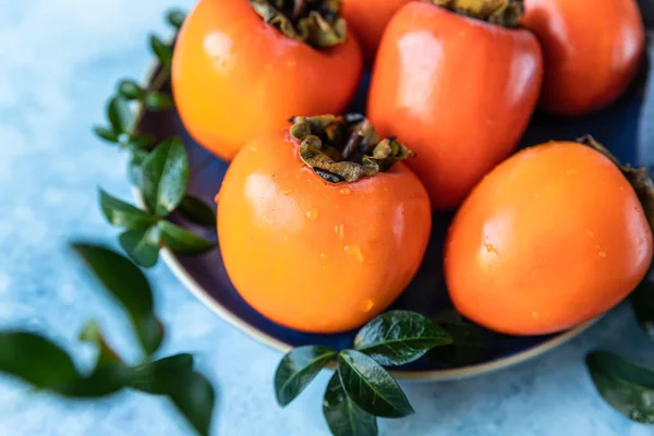 Délicieux fruits mûrs de kaki aux feuilles vertes sur fond de béton bleu. — Photo