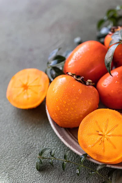 Whole and cut persimmon fruit with water drops and green leaves on green concrete background. — Stock Photo, Image