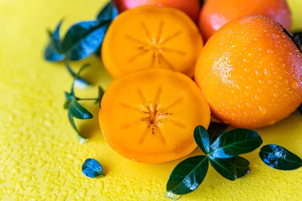 Whole and cut half persimmons with leaves and water drops on yellow background. — Stock Photo, Image