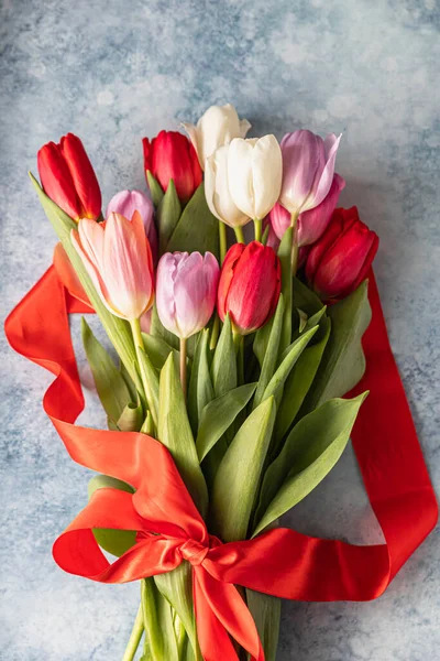 Bukiet kolorowych tulipanów z czerwoną wstążką na niebieskim tle betonowym. Piękne tulipany na Dzień Matki lub Wielkanoc. — Zdjęcie stockowe