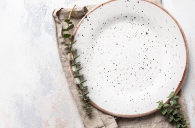 Boş beyaz tabak, keten peçete ve okaliptüs dalları, açık beton arka plan. Bahar menüsü kavramı. Moda minimal sofra takımı. Restoran için model menü. Üst görünüm, alanı kopyala.