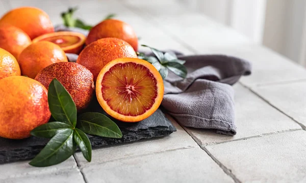 Sliced and whole ripe juicy Sicilian blood oranges with leaves on black stone board, grey tiled background. Seasonal fruits.