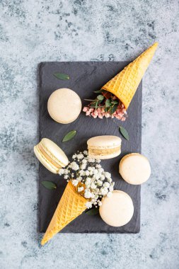 Gri arka planda yapraklı badem kurabiyesi ve çigila çiçekleriyle dolu waffle külahı. Pastel renkler. Geleneksel Fransız badem tatlısı, tatlı dolgulu. Üst görünüm.