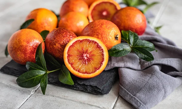 Sliced and whole ripe juicy Sicilian blood oranges with leaves on black board, grey tiled background. Seasonal fruits.