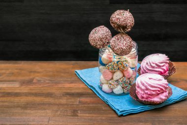 Lezzetli çikolatalı kek, renkli şekerlerle süslenmiş kavanozda beyaz şekerler, peçetede pembe şekerlemeler, fotokopi alanı olan ahşap arka plan, pastalar, çubuk üzerinde tatlılar..