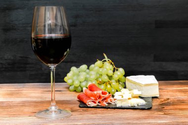 Bardakta kırmızı şarap, brie peyniri, mavi peynir, üzümler, jamon eti, siyah tahtada salam, ahşap masada ahşap masada, atıştırmalıklarla alkol içeceği, hayat yakın..