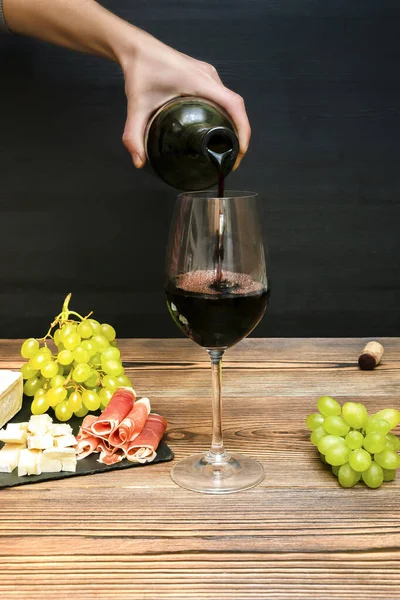 Woman hand pouring Red wine in glass from bottle, brie cheese, blue cheese, grapes, jamon meat, salami on black slate board on wooden table, alcohol drink with snacks, still life.