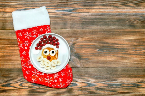 Christmas Santa Claus face shaped pancake with sweet fresh raspberry berry, banana on plate, christmas sock on wooden background for kids children breakfast. xmas food dessert, new year decorations — Stockfoto