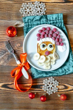 Noel Baba, çocukların kahvaltısı için ahşap arka planda tatlı taze ahududu ve muzlu krep şeklinde bir pankek sunar. Noel tatlısı, yeni yıl süslemeleri, kırmızı toplar.