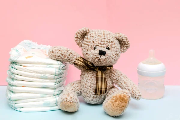 Stack of diapers with cute teddy bear toy, bottle of milk on table. set for infant boy girl for baby shower present gift on pink blue background. Healthcare medical, hygiene concept — Stockfoto