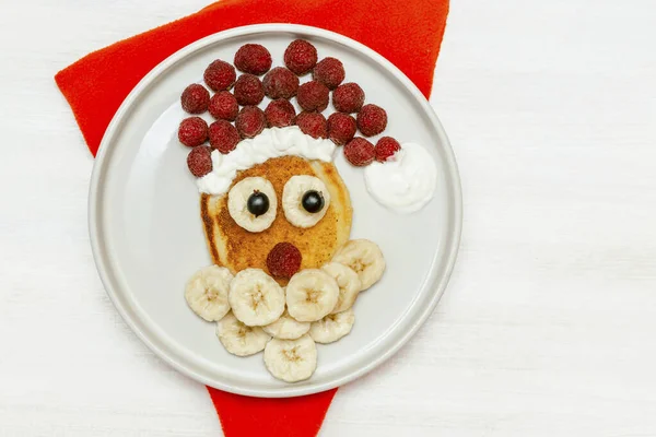 Christmas Santa Claus face shaped pancake with sweet fresh raspberry berry and banana on plate, red napkin on white wooden background for kids children breakfast. xmas sugar food dessert — Stockfoto