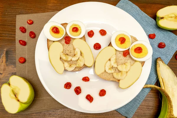 Funny Hibou visage pain grillé avec des œufs de poulet, pomme, banane, fruits secs de baie dans l'assiette. Enfants mignons enfants bébés dessert sucré petit déjeuner, déjeuner, art alimentaire sur fond en bois, vue sur le dessus — Photo