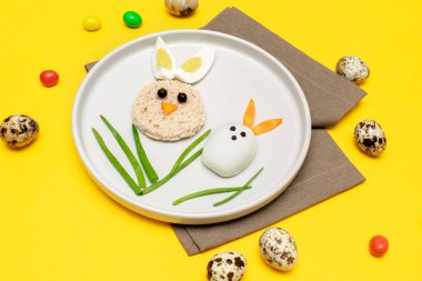 Çocuklar ve çocuklar için Paskalya komik, yaratıcı, sağlıklı öğle yemeği fikri. Tavşan, haşlanmış tavuk yumurtasından, ekmekten, soyulmuş havuçtan, yeşil tabakta sarı masa zemininde..