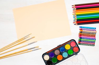 Suluboya boyaları, boya fırçaları, renkli kalemler, pastel boya kalemleri, boş suluboya kağıt pedler. Yaratıcılık yaratma süreci. Sanatçının eşyaları beyaz masada. Çizim malzemelerinin üst görünümü.
