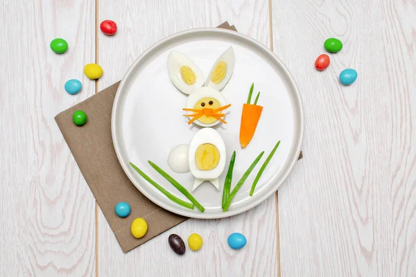 Easter funny creative healthy breakfast lunch food idea for kids, children. Bunny, rabbit made from boiled chicken eggs,peeled carrots, greens on plate white wood table background. Top view Flat lay.