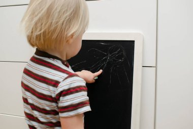 Küçük sevimli beyaz çocuk evdeki tahtaya tebeşir çiziyor. Bebek eli boyama. Çocuklar için yaratıcı kapalı alan aktivitesi, gelişim, çocuk yuvası, anaokulu konsepti.