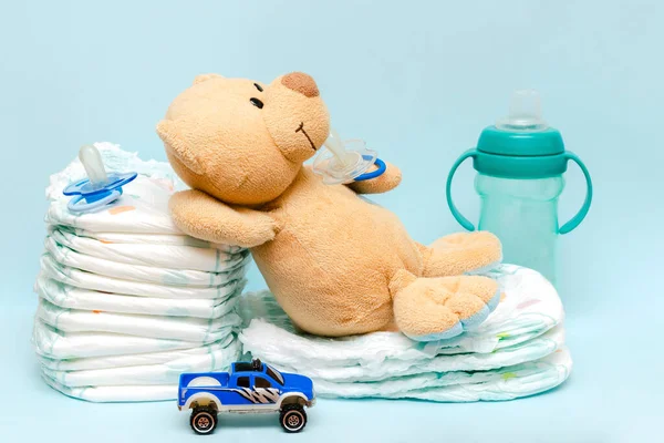 Stack of diapers with cute teddy bear car toy pacifier bottle lying on table. set for infant newborn boy girl for baby shower present gift on blue background. Healthcare medical, hygiene concept.