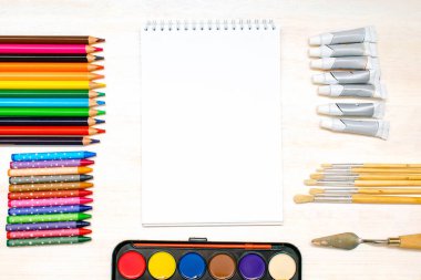 Suluboya boyaları, boya fırçaları, renkli kalemler, pastel boya kalemleri, boş suluboya kağıt pedler. Yaratıcılık yaratma süreci. Sanatçının eserleri beyaz masada. Resim malzemelerinin üst görünümü..