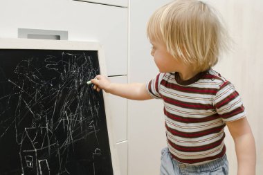Küçük sevimli beyaz çocuk evdeki tahtaya tebeşir çiziyor. Bebek eli boyama. Çocuklar için yaratıcı kapalı alan aktivitesi, gelişim, çocuk yuvası, anaokulu konsepti.