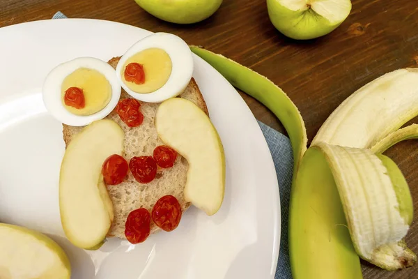 Funny owl face sandwich toast bread with chicken eggs, apple, banana, dried berry fruits on plate. Cute kids childrens baby\'s sweet dessert breakfast,lunch, food art on wooden background,top view.