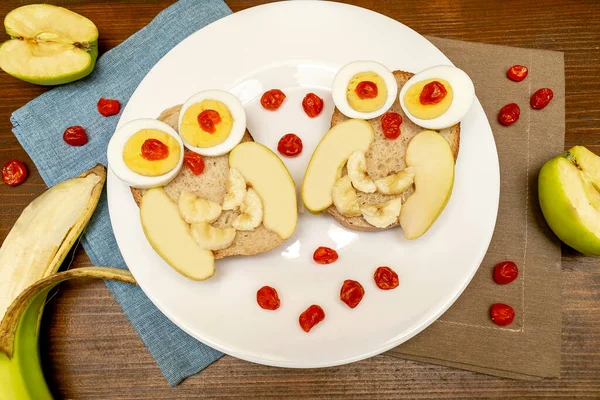 Funny owl face sandwich toast bread with chicken eggs, apple, banana, dried berry fruits on plate. Cute kids childrens baby\'s sweet dessert breakfast,lunch, food art on wooden background,top view.