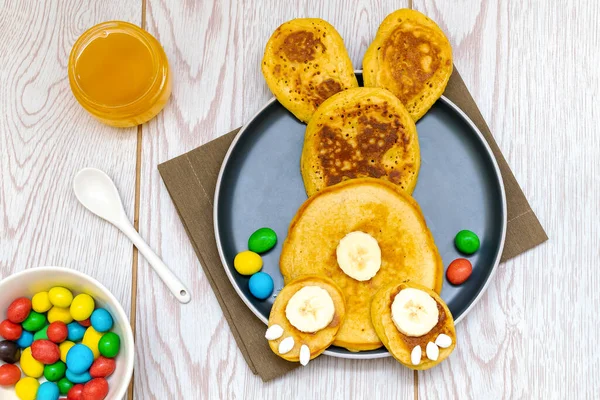 Easter funny creative healthy breakfast lunch food idea for kids, children.Bunny, rabbit made from pancakes,banana with sweet candies,honey on plate wooden table background.Top view Flat lay.