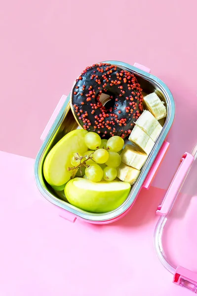 Catering food prepared in storage container with compartment with doughnut,apple,banana,cookies on pink background. meal with healthy balanced diet, lunch box boxed take away delivery packed ready.
