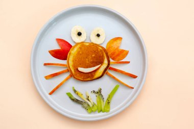 Krep, elma, muz, kivi tabağından komik yengeç suratlı atıştırmalık. Sevimli çocuklar bebeğin tatlı tatlısını, sağlıklı kahvaltısını, öğle yemeğini, bej arka planda yemek sanatını, en üstteki düz manzarayı....