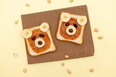 Funny cute bear faces sandwich toast bread with peanut butter, banana,nuts. Kids childrens baby's sweet dessert healthy breakfast lunch food art on plate on yellow background,close up,top view.