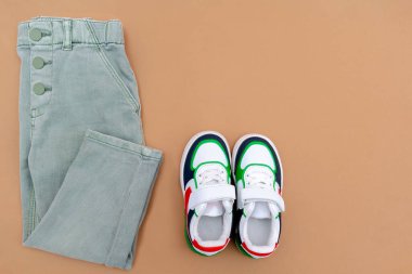 Bebek ayakkabısı ve kot pantolon. Bahar, sonbahar ya da yaz için çocuk kıyafetleri ve aksesuarlar. Moda çocuk kıyafetleri, bej kahverengi arka planda giysiler. Düz yatıyordu, üstten, yukarıdan..