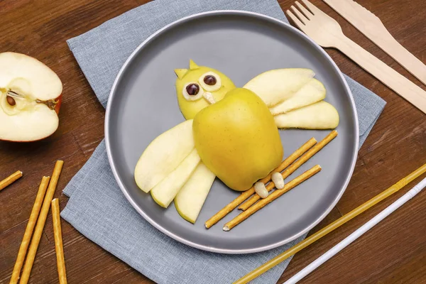 Funny owl face shape snack with apple, wheat crispy bread straw on plate. Cute kids childrens baby's sweet dessert, healthy breakfast,lunch, food art on wooden background,top view.