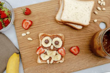 Komik baykuş suratlı tost ekmeği, fıstık ezmesi, muz, ahşap tahtada çilek. Tatlı çocuklar bebeğin tatlı tatlısını, sağlıklı kahvaltısını, öğle yemeğini, gri arka planda yemek sanatını, üst manzarayı....