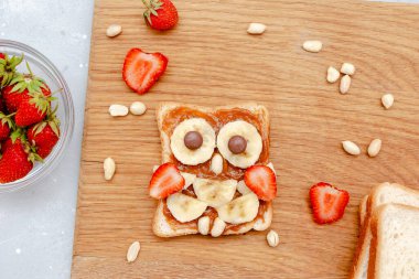Komik baykuş suratlı tost ekmeği, fıstık ezmesi, muz, ahşap tahtada çilek. Tatlı çocuklar bebeğin tatlı tatlısını, sağlıklı kahvaltısını, öğle yemeğini, gri arka planda yemek sanatını, üst manzarayı....