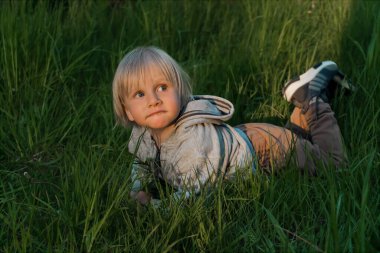 Küçük sevimli beyaz çocuk bahçedeki çimlerin üzerinde uzanırken eğleniyor. Gün batımında bahçede çimenlikte bir çocuk. Mutlu çocukluk, çocuk sağlığı, huzur konsepti..
