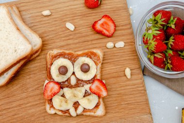 Komik baykuş suratlı tost ekmeği, fıstık ezmesi, muz, ahşap tahtada çilek. Tatlı çocuklar bebeğin tatlı tatlısını, sağlıklı kahvaltısını, öğle yemeğini, gri arka planda yemek sanatını, üst manzarayı....