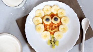 Funny chamomile flower face shape snack from pancake,bananas,kiwi.Cute kids childrens baby's sweet dessert,healthy breakfast,lunch,food art with milk on plate,gray concrete background,top view.