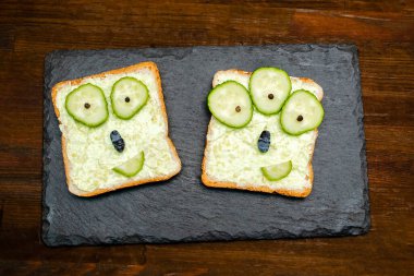Cadılar bayramı sandviçinde komik canavar suratları yumuşak yeşil avokado peynirli ricotta tereyağlı ekmek servis tahtasında salatalık, tahta zeminde yarasalar. Çocuklar çocuk kahvaltısı öğle yemeği.