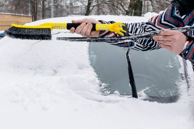 Adam arabanın ön camından fırçayla kar ve buz fırçalıyor. Erkek işçi kar fırtınasından sonra taze kar tabakasını temizliyor, kışın ön camdan kar yağıyor..