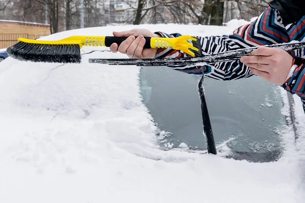 Adam arabanın ön camından fırçayla kar ve buz fırçalıyor. Erkek işçi kar fırtınasından sonra taze kar tabakasını temizliyor, kışın ön camdan kar yağıyor..