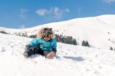 Little cute baby kid child boy in winter overalls,ski mask,glasses goggles sitting on snow at winter holiday, in ski resort in Carpathian high mountains outdoor nature landscape, Ukraine, Europe.