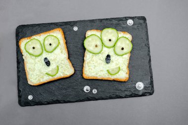 Funny monster faces on halloween sandwich toast bread with soft green avocado cheese ricotta butter,cucumbers on cutting serving board,bats on gray background.Kids child breakfast lunch food.