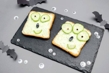 Funny monster faces on halloween sandwich toast bread with soft green avocado cheese ricotta butter,cucumbers on cutting serving board,bats on gray background.Kids child breakfast lunch food.