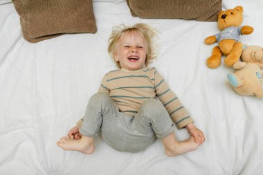 Küçük sevimli sarışın beyaz çocuk oyun oynuyor, eğleniyor, yatağa uzanıyor, güneşli bir sabah uyanıp odasında bacaklarını kaldırıyor. Mutlu çocukluk anları.