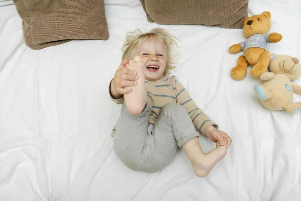 Cute little blond caucasian child kid toddler boy playing,having fun, lying on bed,lifts up the legs at home at sunny morning wakes up in his room. happy childhood moments.