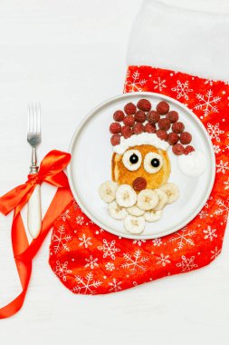 Christmas Santa Claus shaped pancake with sweet fresh raspberry berry and banana on plate, red sock, fork on white wooden background for kids children breakfast. xmas food with new year decorations.